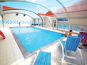 Ein Hallenbad mit klarem Wasser, umgeben von Liegestühlen und einem überdachten Bereich mit Schachbrettmuster-Wand.