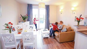 A modern living room with a sofa, dining table, and chairs. There is a kitchen area with a counter and a window. - Relaxing Coastal Apt for 8 (le Crotoy)