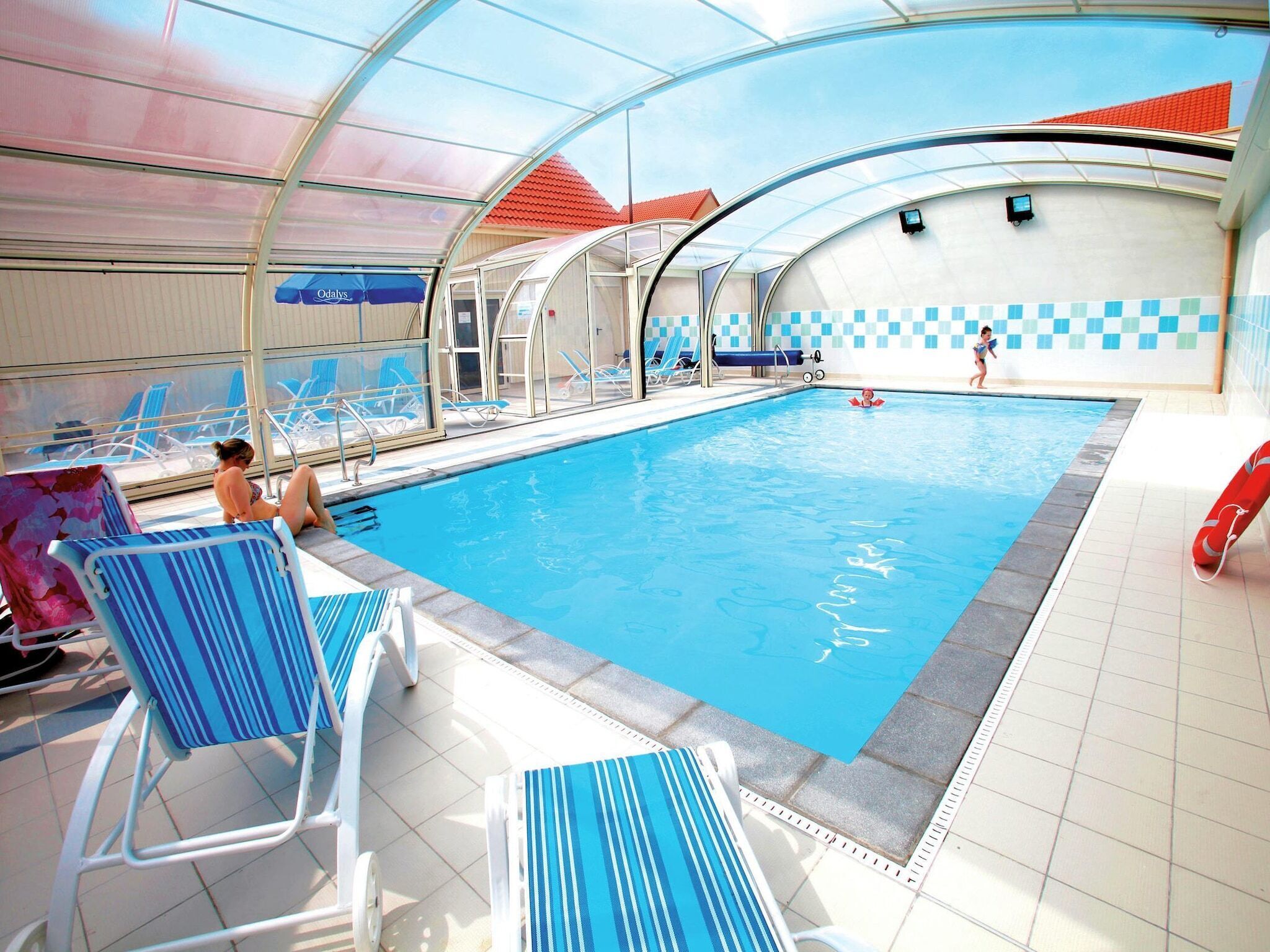 An indoor swimming pool with clear water, surrounded by blue-striped lounge chairs and a covered area with a red roof.