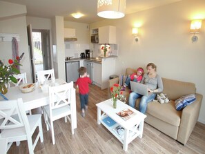 A modern living room with a sofa, a dining table with chairs, a kitchen area with a dishwasher, and a small table with a vase of flowers.