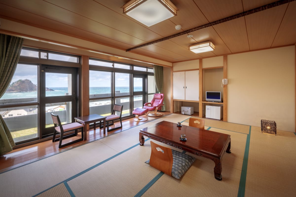 Een kamer met tatami vloer, een houten bureau, een rode stoel, een televisie en uitzicht op zee.