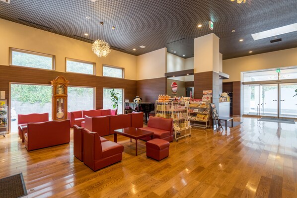 A spacious lobby with wooden flooring, red seating, a large clock, and a reception area with a counter and informational posters. - Hotel Sunresort Shonai (Tsuruoka)