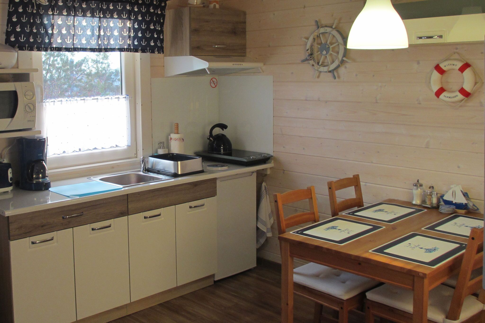 Une cuisine compacte équipée d’un four à micro-ondes, d’une bouilloire et d’un évier. On y trouve une table en bois avec des chaises, et une décoration murale représentant un gilet de sauvetage.