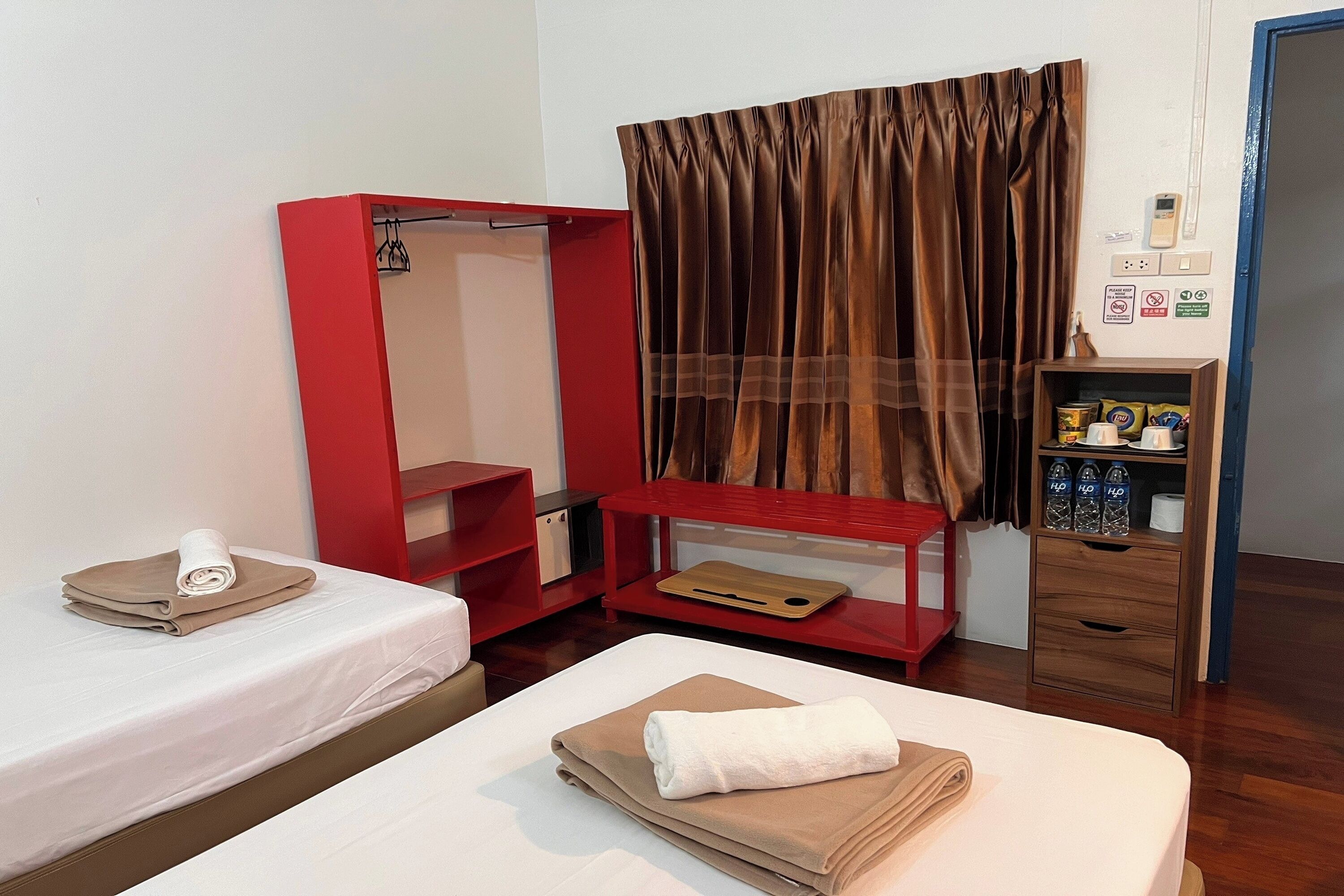 A hotel room with two beds, a red wardrobe, a wooden shelf with toiletries, and a curtain.