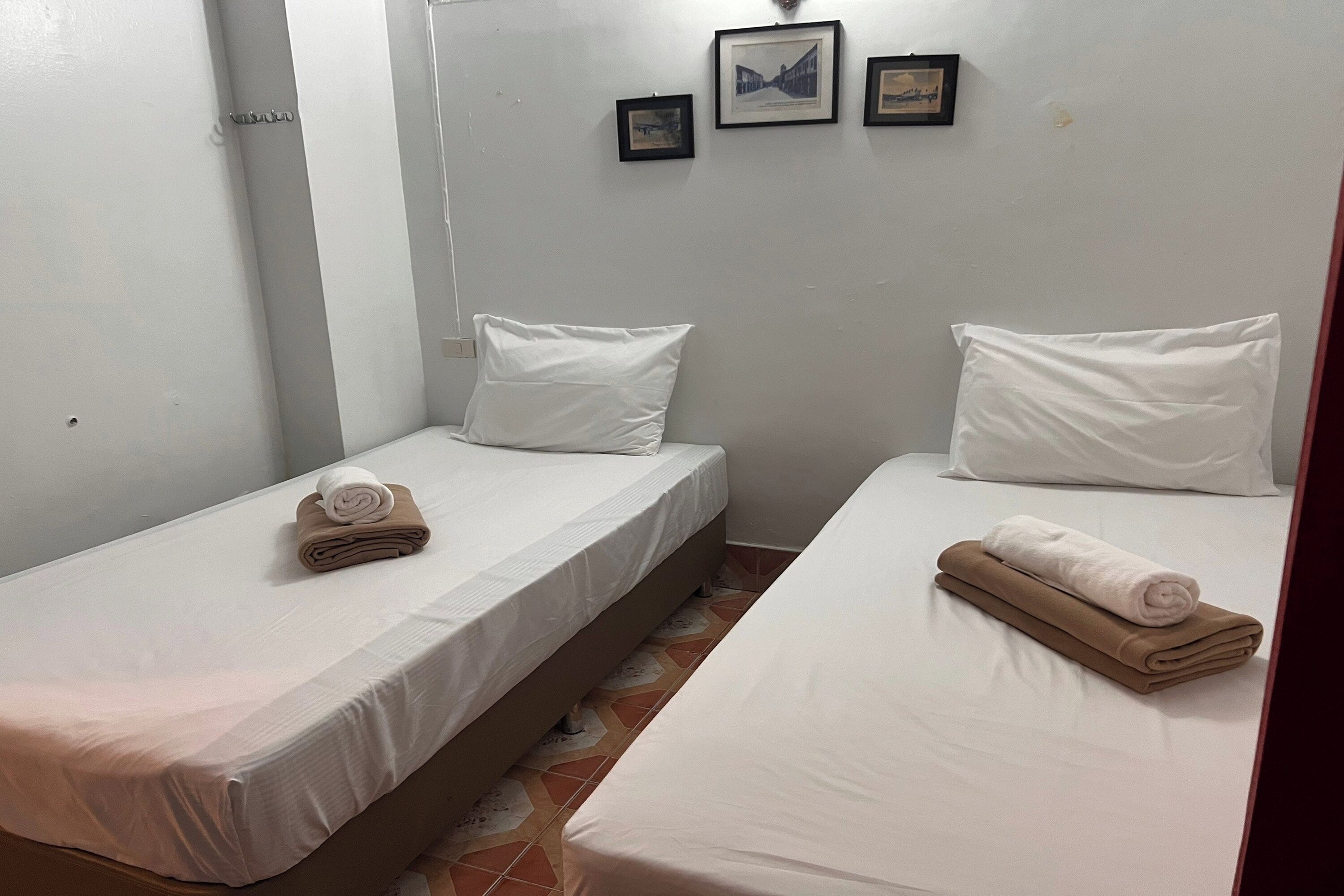Deux lits simples avec du linge de lit blanc et des serviettes marron, un sol carrelé et un mur orné de trois tableaux encadrés.