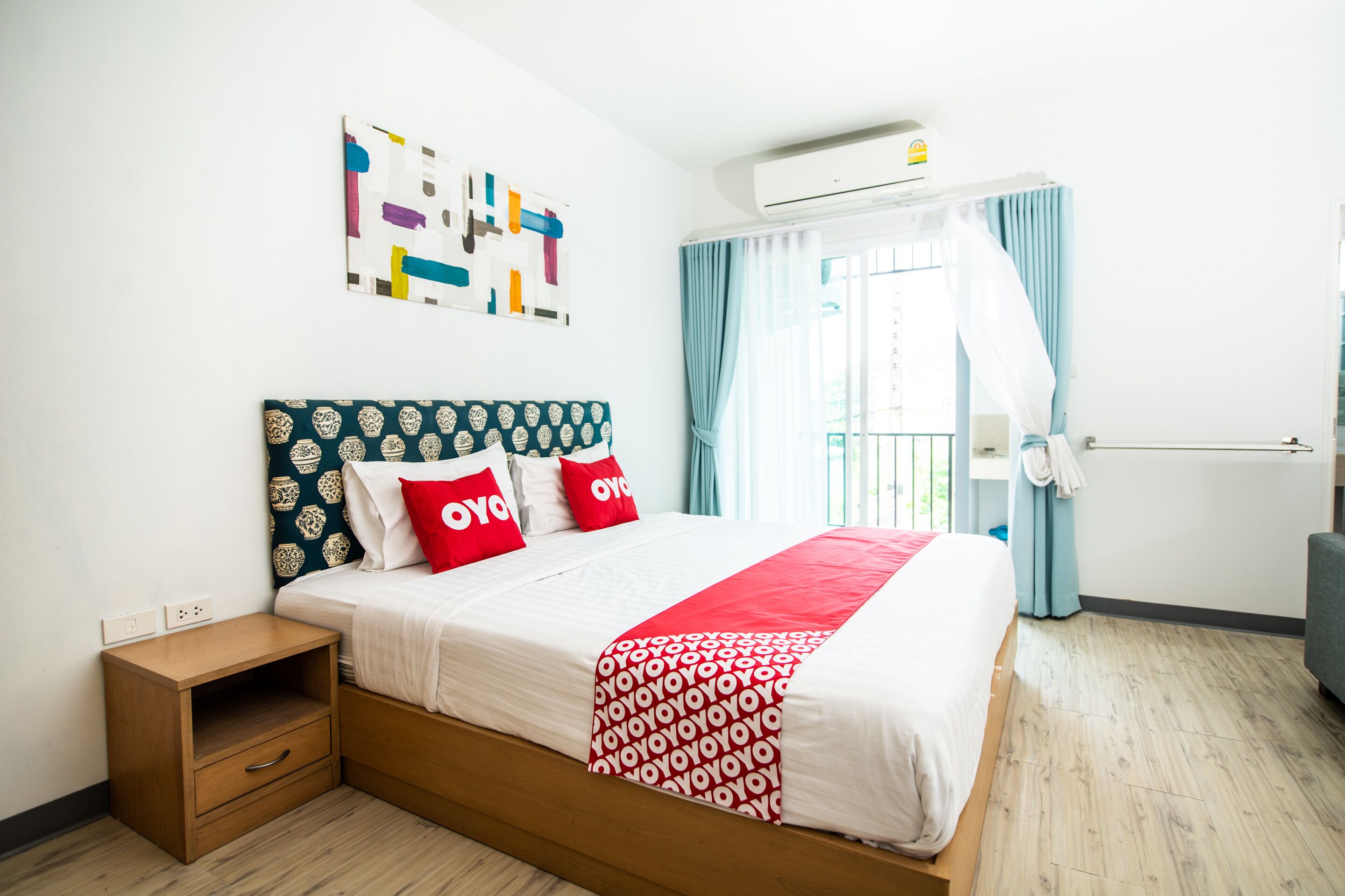 A neatly arranged hotel room with a bed, a small wooden nightstand, a wall-mounted air conditioner, and a window with curtains.