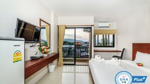 A hotel room with a bed, a desk with a TV, a window with curtains, a balcony, and an air conditioning unit. - The Topaz Residence (Phuket)