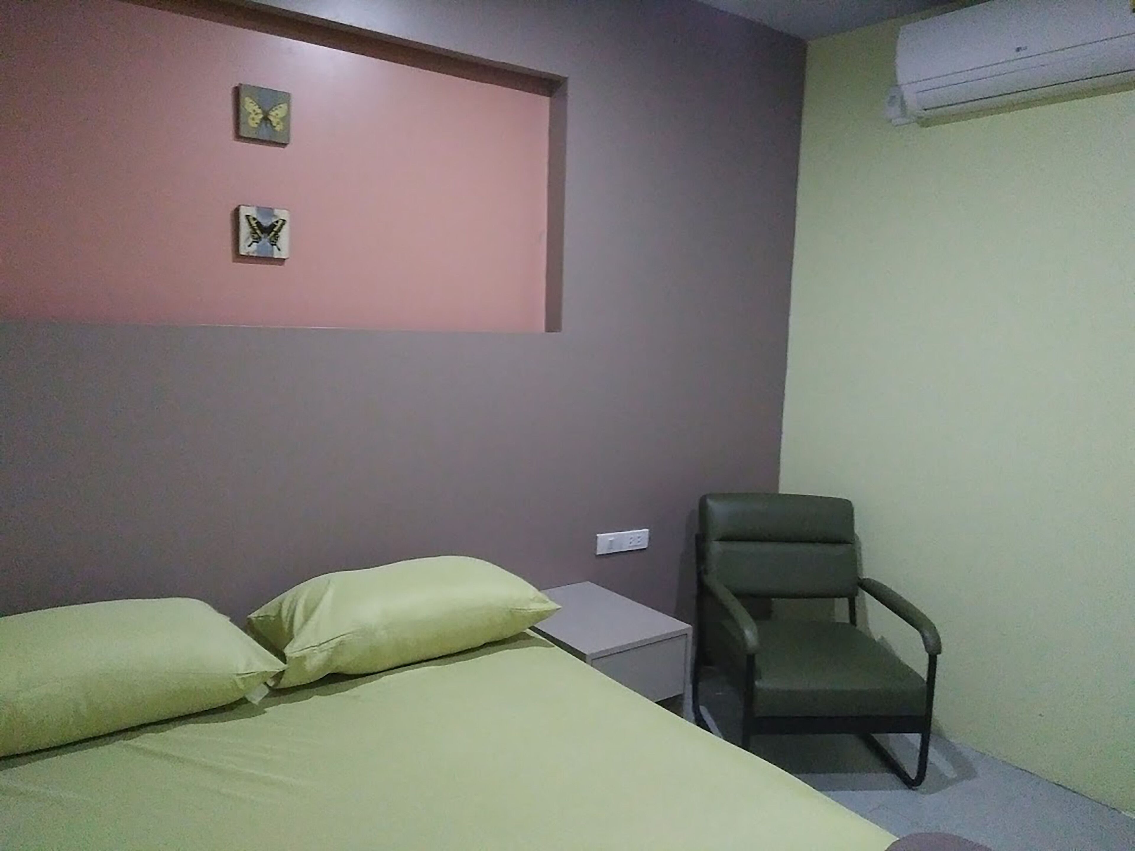 A room with a bed, two pillows, a chair, a small table, and an air conditioning unit.