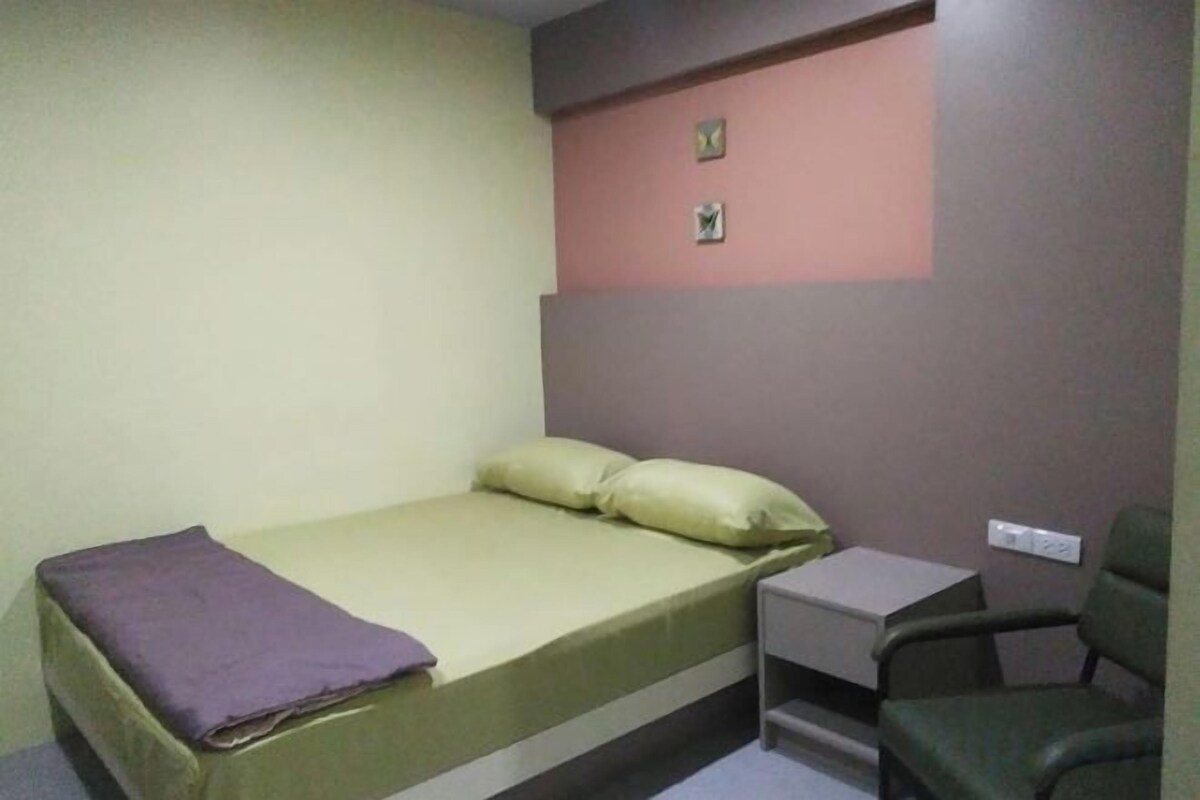 Una cama individual con colchón y almohadas, una mesita de noche y una silla en una habitación con paredes lisas.