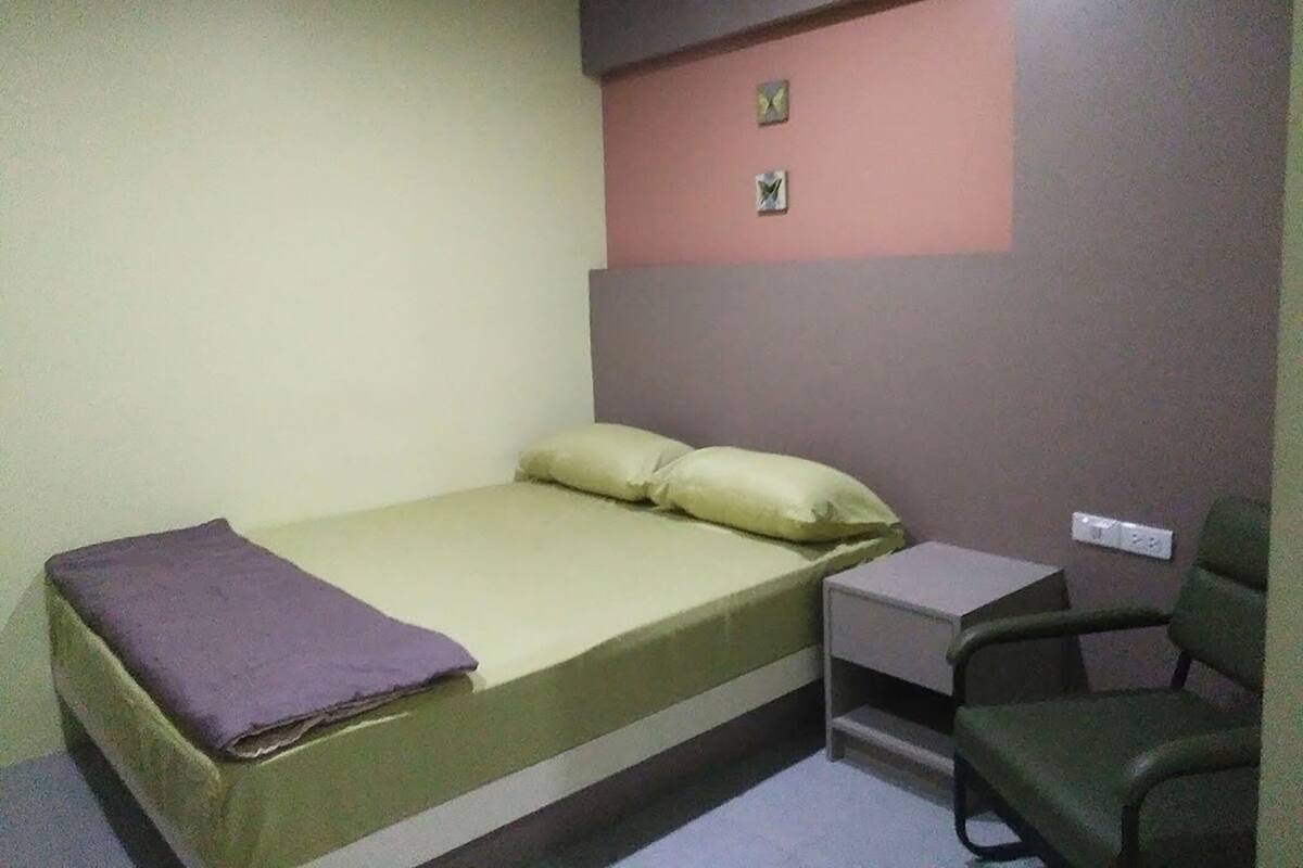 Una habitación pequeña con cama individual, una cama verde, una manta morada, una silla verde y un escritorio pequeño con silla.