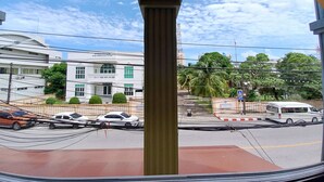 Une vue à travers une fenêtre en arc d’un bâtiment à la façade blanche et aux encadrements de fenêtres verts, une rue avec des voitures et un autobus, et un ciel dégagé avec des nuages.