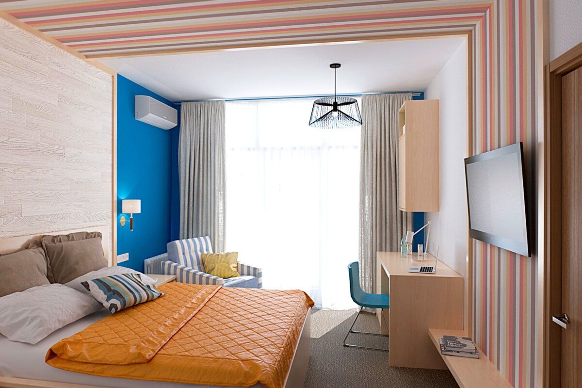 A hotel room with a bed, a desk, a chair, a TV, and a window with curtains.