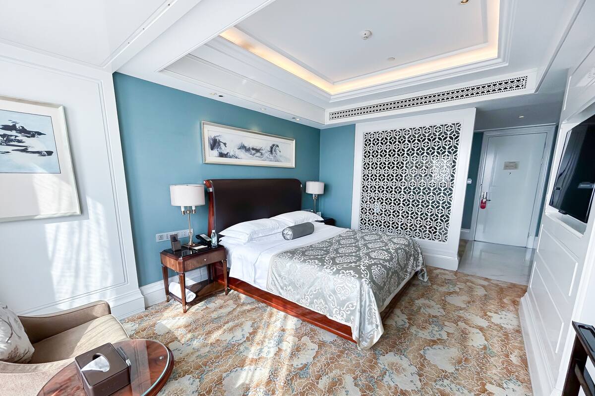 Une chambre d’hôtel avec un lit, des tables de chevet, une lampe, une chaise, une table et un tapis à motifs.
