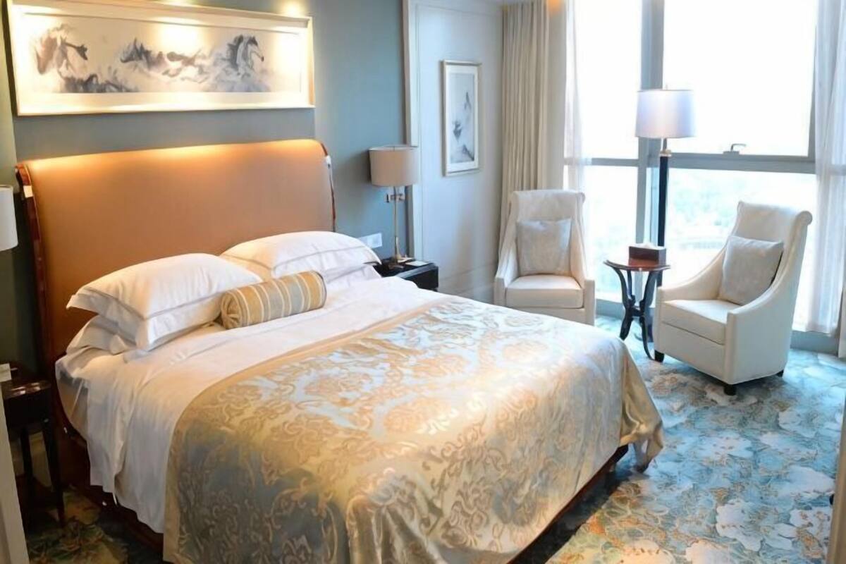 Une chambre d’hôtel avec un grand lit, deux chaises, une petite table et une fenêtre avec des rideaux.