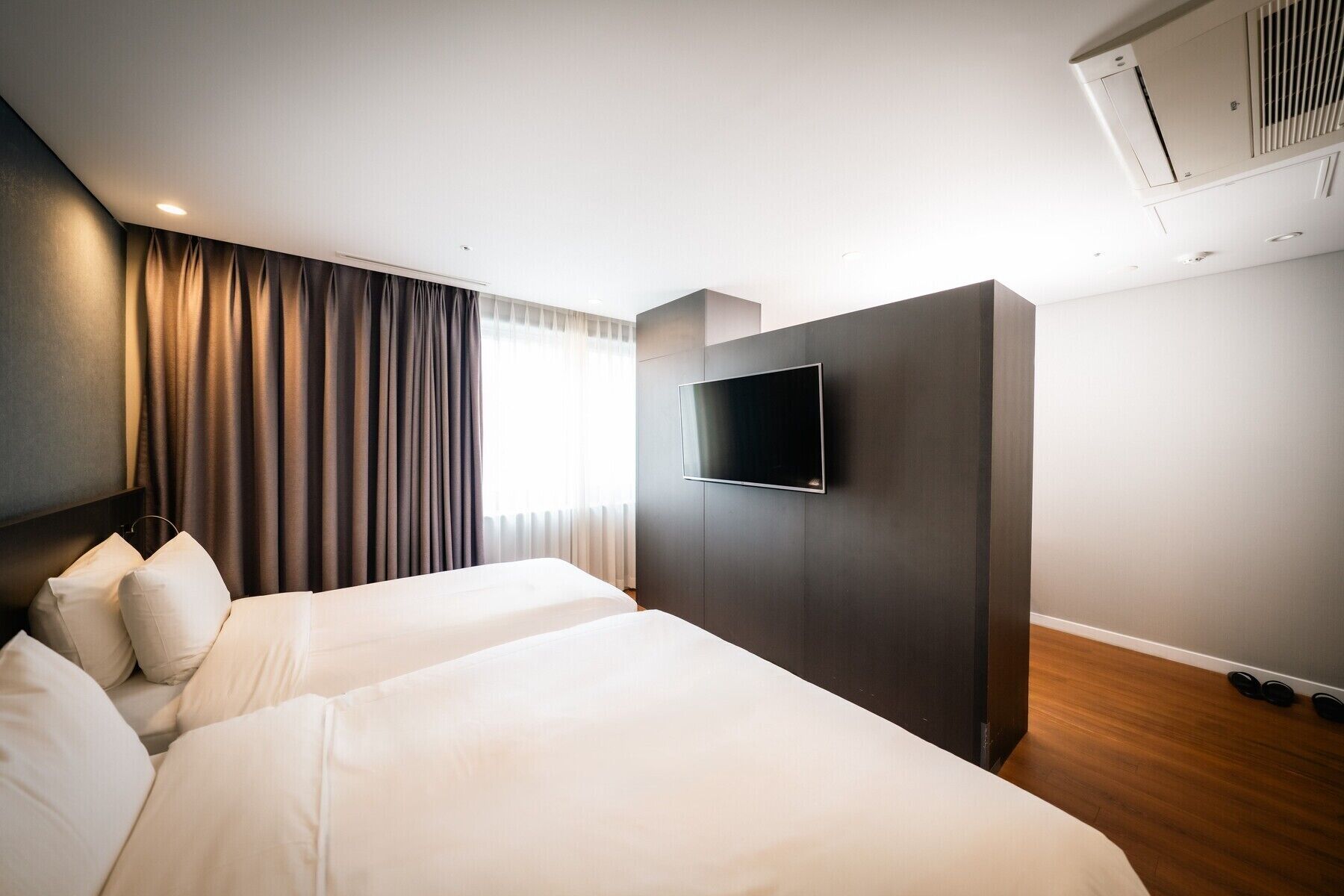 A hotel room with two beds, a flat-screen TV mounted on the wall, and a large window with curtains.