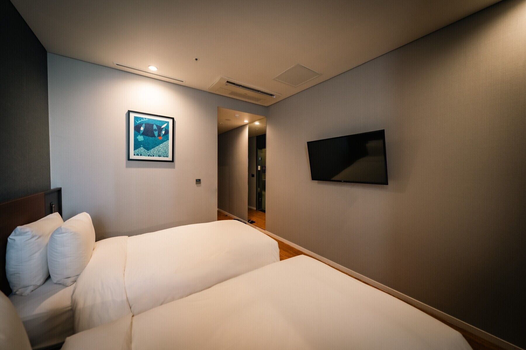 Ein Hotelzimmer mit zwei Betten, einem an der Wand montierten Flachbildfernseher und einem gerahmten Kunstwerk über dem Bett links.