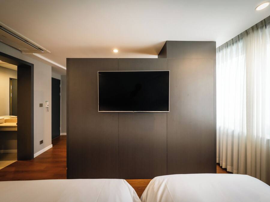 Una habitación de hotel moderna con un televisor de pantalla plana montado en una pared oscura, suelo de madera y un gran ventanal con cortinas traslúcidas.