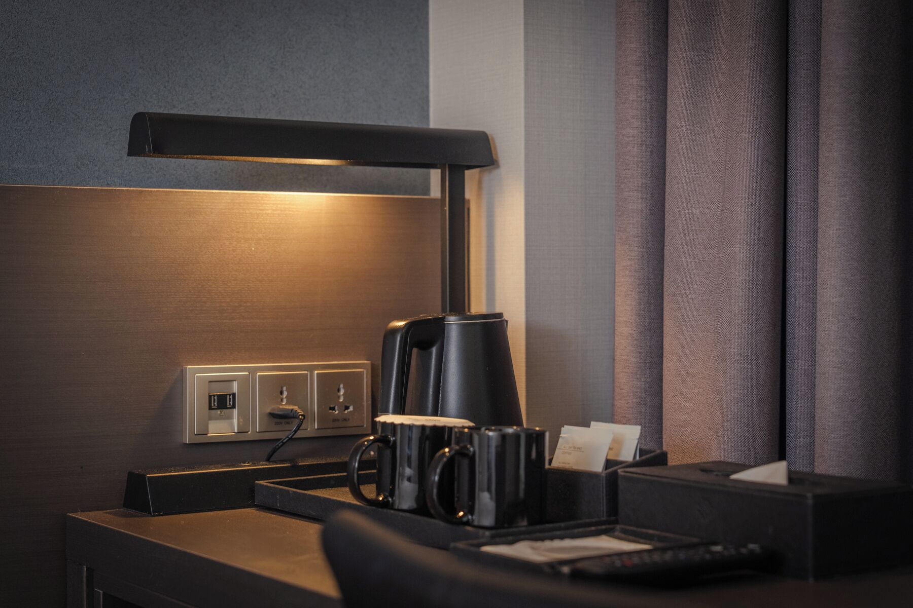 A hotel room with a desk, a coffee maker, a lamp, and a power outlet.