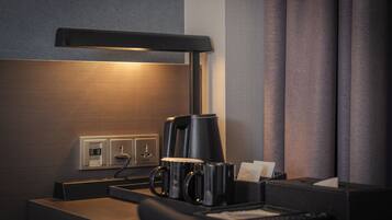 A hotel room with a desk, a coffee maker, a lamp, and a power outlet.