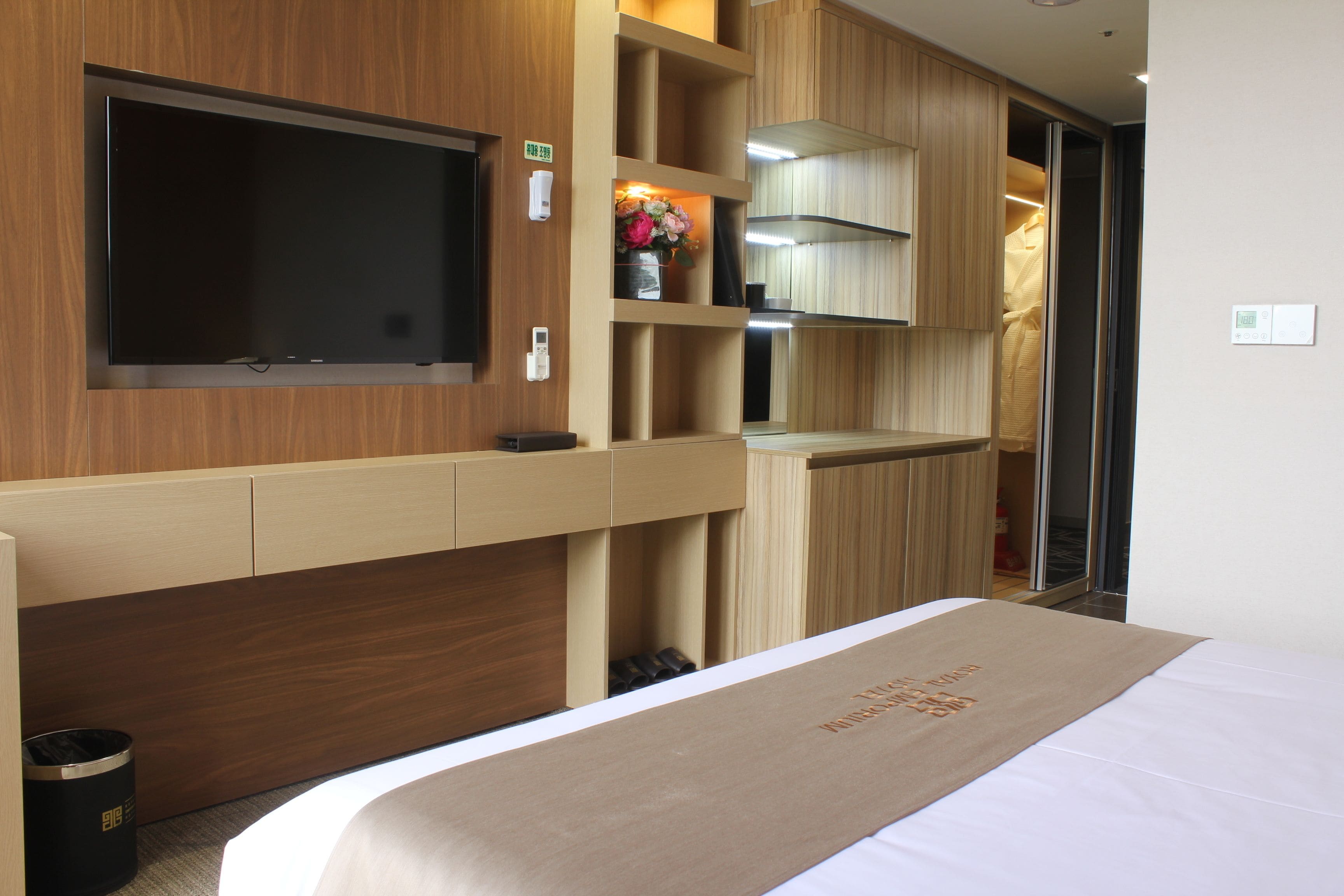Ein Hotelzimmer mit Bett, einem an der Wand montierten Fernseher, einem Holzschrank und einer kleinen Ablage mit Blumen in einer Vase.