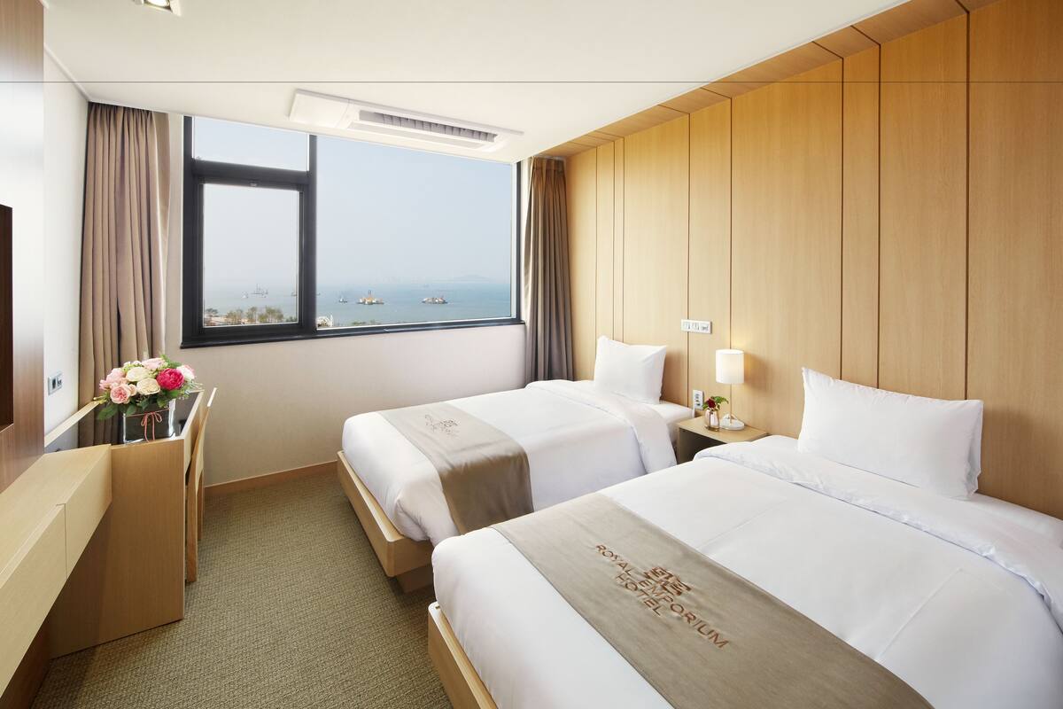 Une chambre d’hôtel avec deux lits, un bureau, une fenêtre donnant sur la mer et un mur recouvert de panneaux en bois.