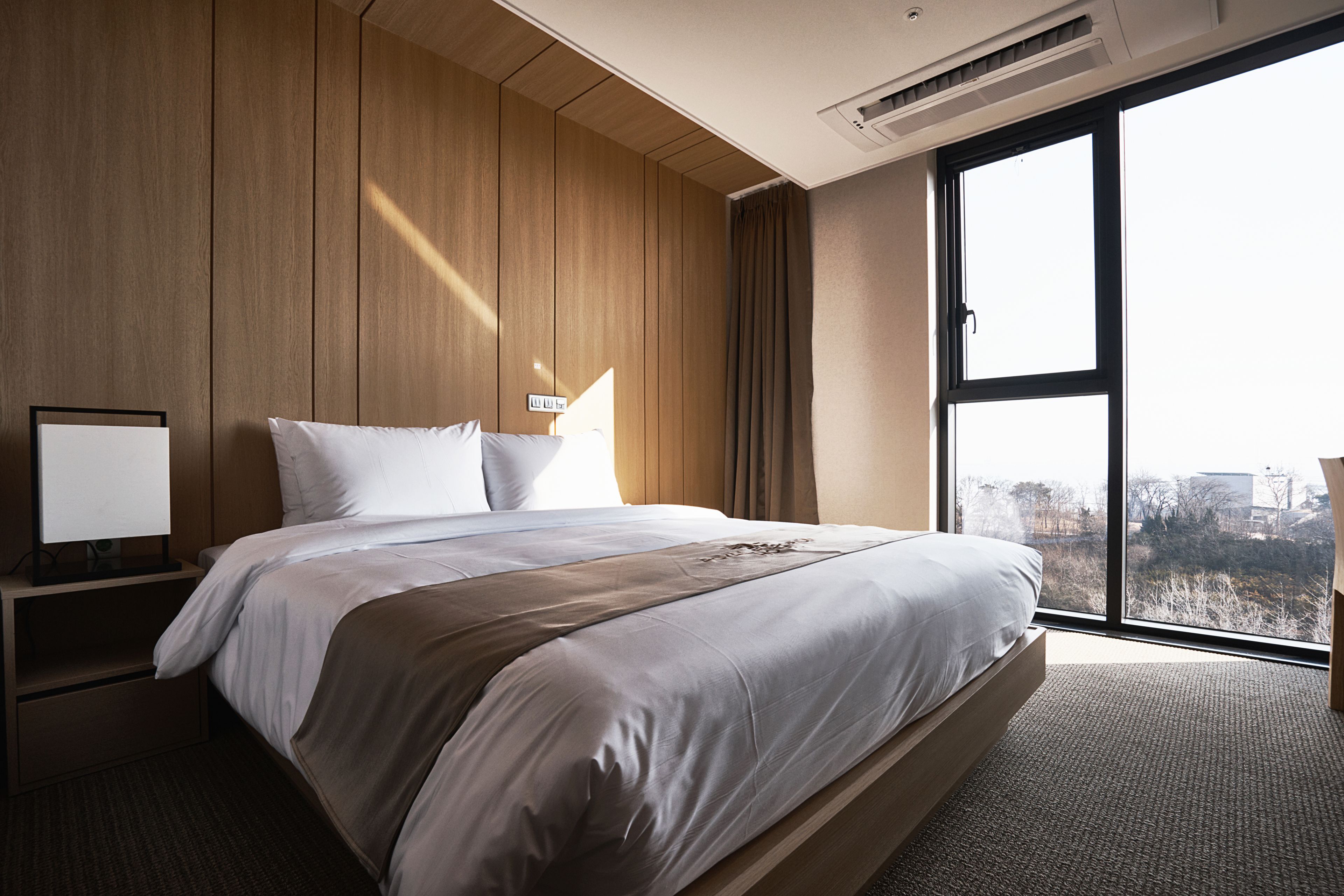 Een moderne hotelkamer met een groot bed, houten paneelwanden, een nachtkastje en een groot raam met uitzicht op de stad.