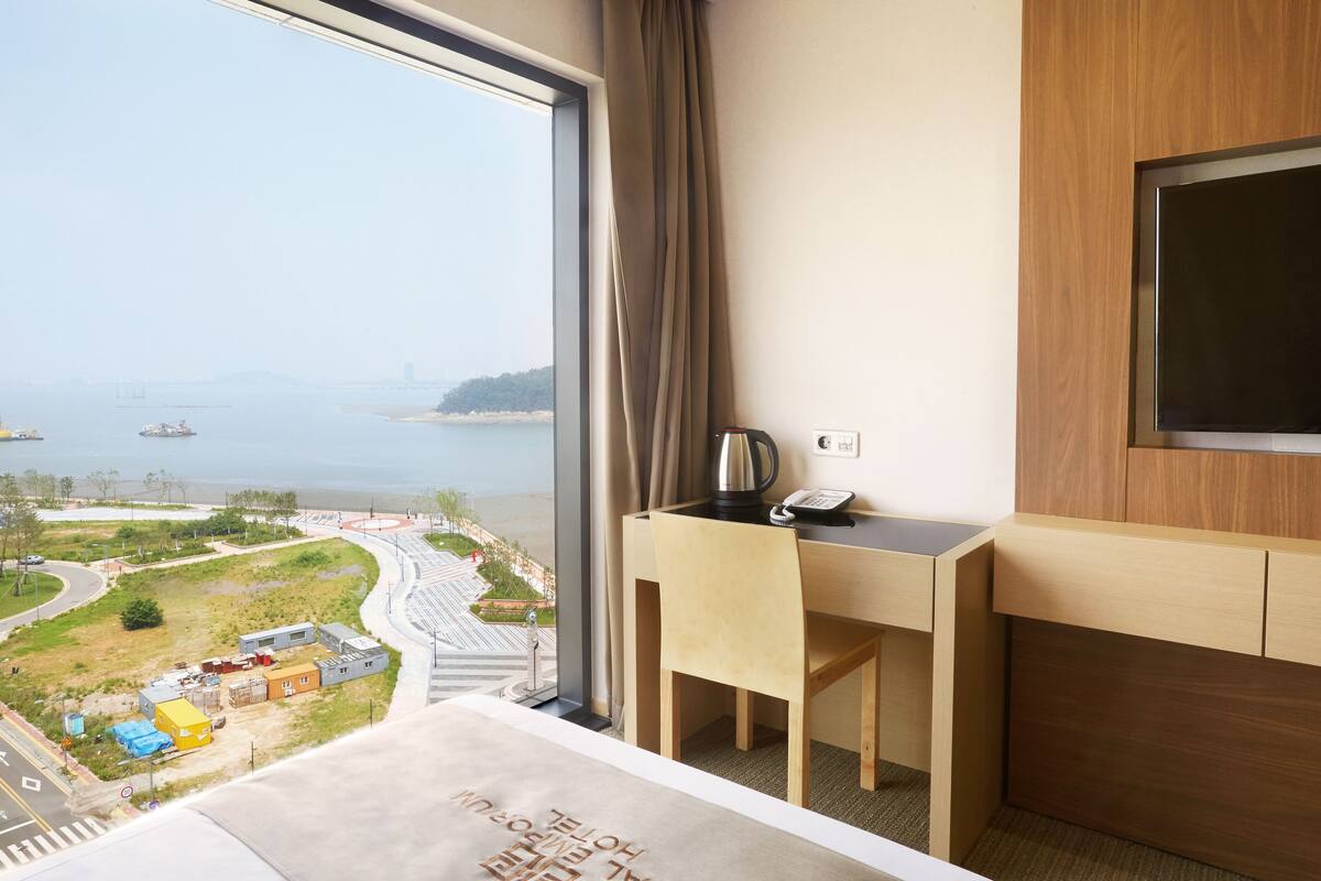 Une chambre d’hôtel dotée d’une grande fenêtre donnant sur la côte, d’un bureau avec une chaise, d’une télévision et d’une bouilloire.