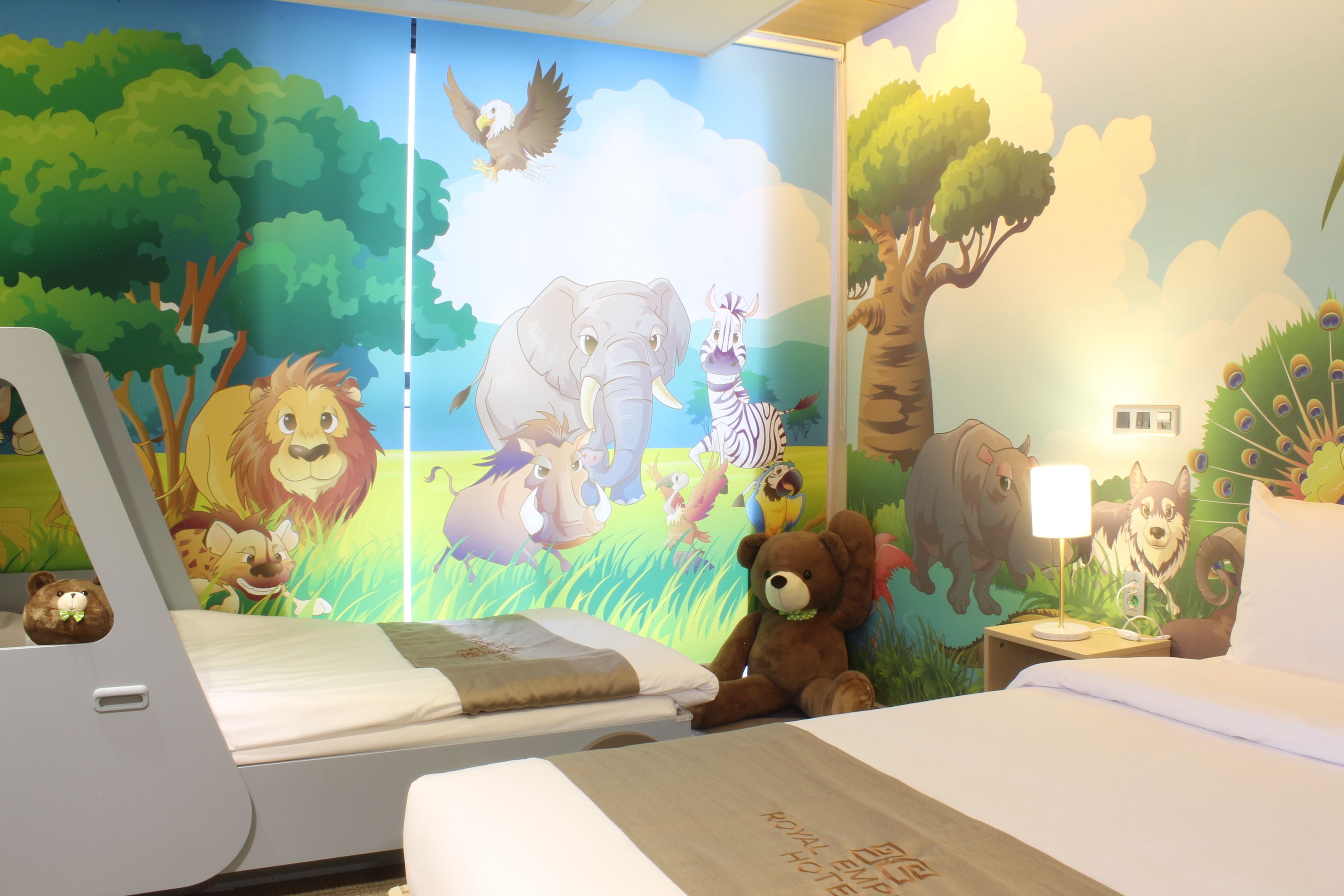 A children's room with a mural of animals, a bed, a teddy bear, and a nightstand with a lamp.