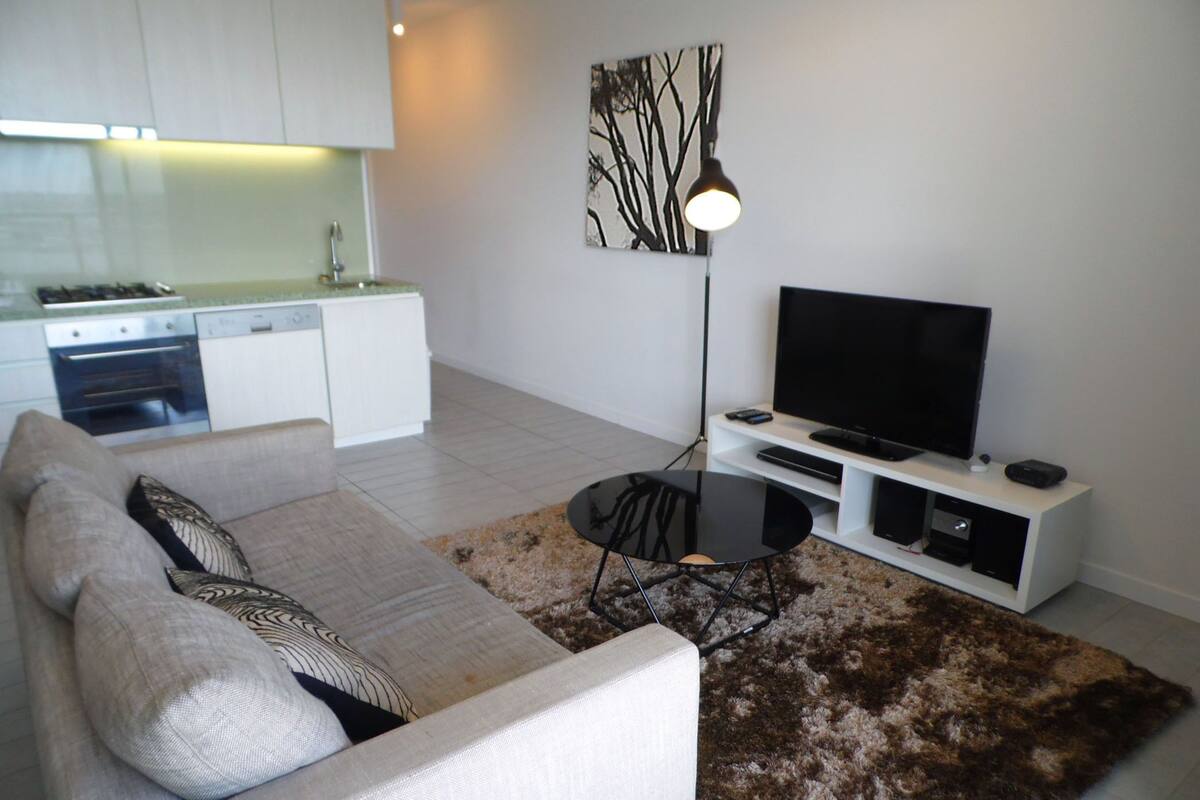 Een moderne woonkamer met een bank, een salontafel, een flatscreen televisie en een keuken op de achtergrond.