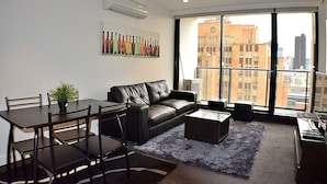 Una sala de estar moderna con un sofá de cuero, una mesa de comedor y sillas. Se aprecia un ventanal con vista a la ciudad, un cuadro con botellas en la pared y un equipo de aire acondicionado.