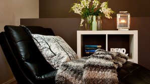 Een zwarte lederen stoel met een kussen met een boommotief en een bont plaid. Achter de stoel staat een witte console tafel met een vaas witte bloemen en een brandende kaars.