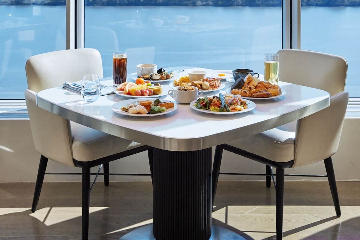 Een eettafel met verschillende gerechten, glazen en een kop, voor een groot raam met uitzicht op de stad.