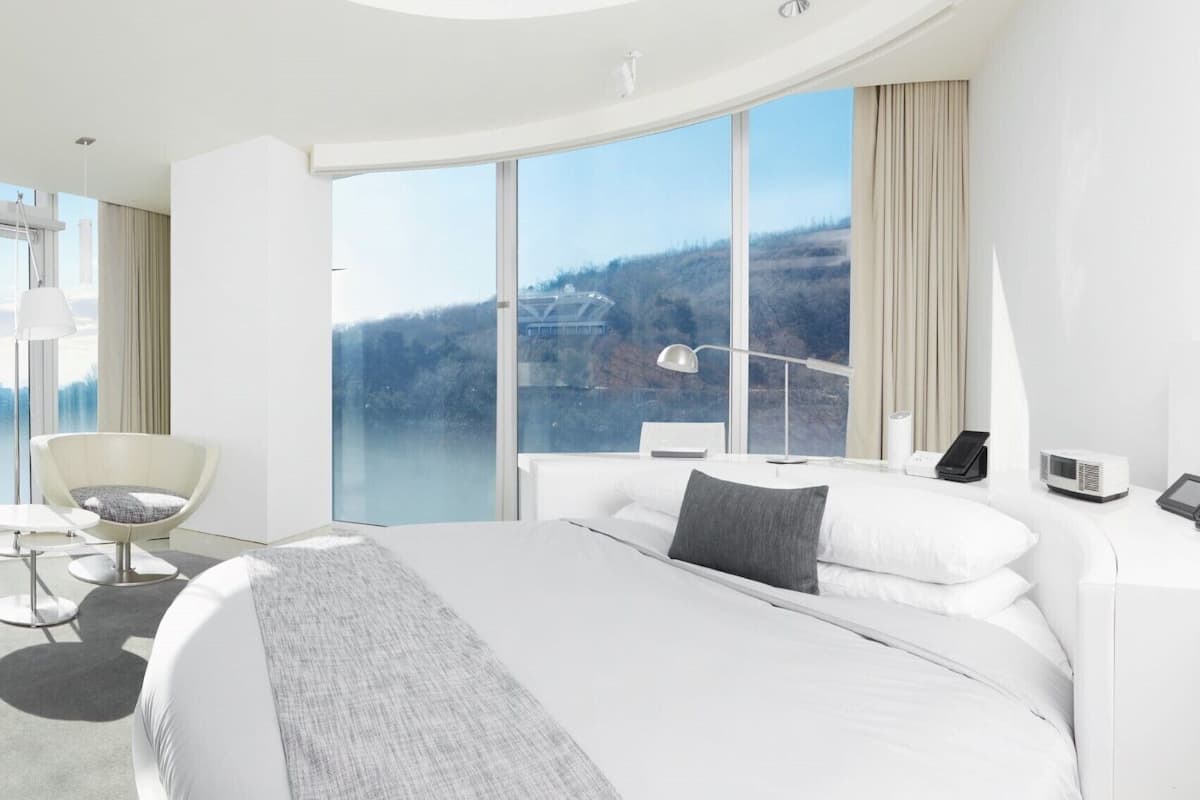 Een moderne hotelkamer met een groot bed, twee fauteuils, een bureau en een uitzicht op een stadsgezicht door grote ramen.