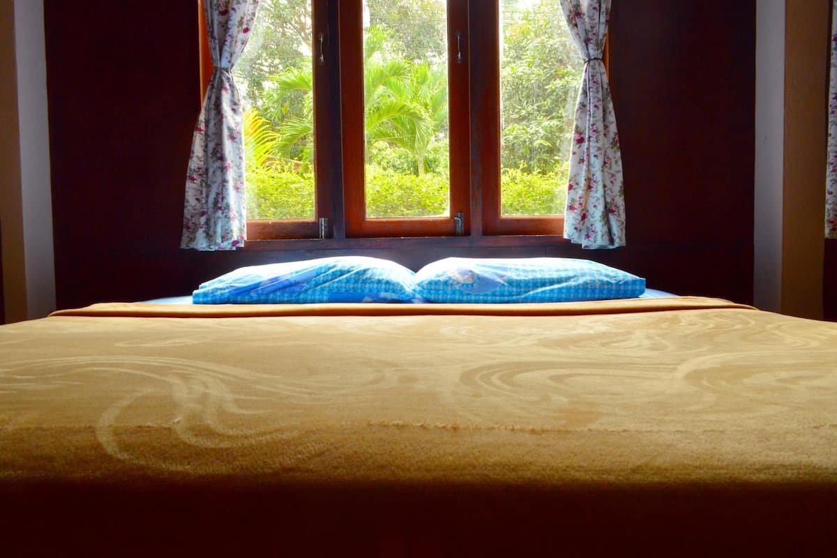 Uma cama com colcha amarela e almofadas azuis, uma janela com cortinas e vista para o verde lá fora.