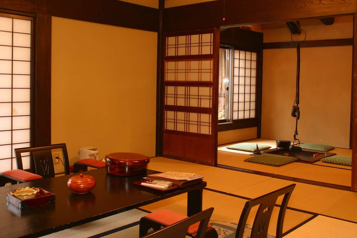 Una habitación japonesa tradicional con suelo de tatami, una mesa baja con un recipiente rojo lacado, una silla de madera y una puerta corredera con diseño de cuadrícula.