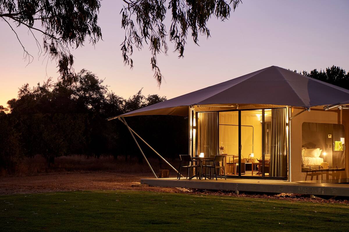 Estrutura moderna e bem iluminada em estilo de tenda, com área de jantar e quarto visíveis através de grandes janelas.