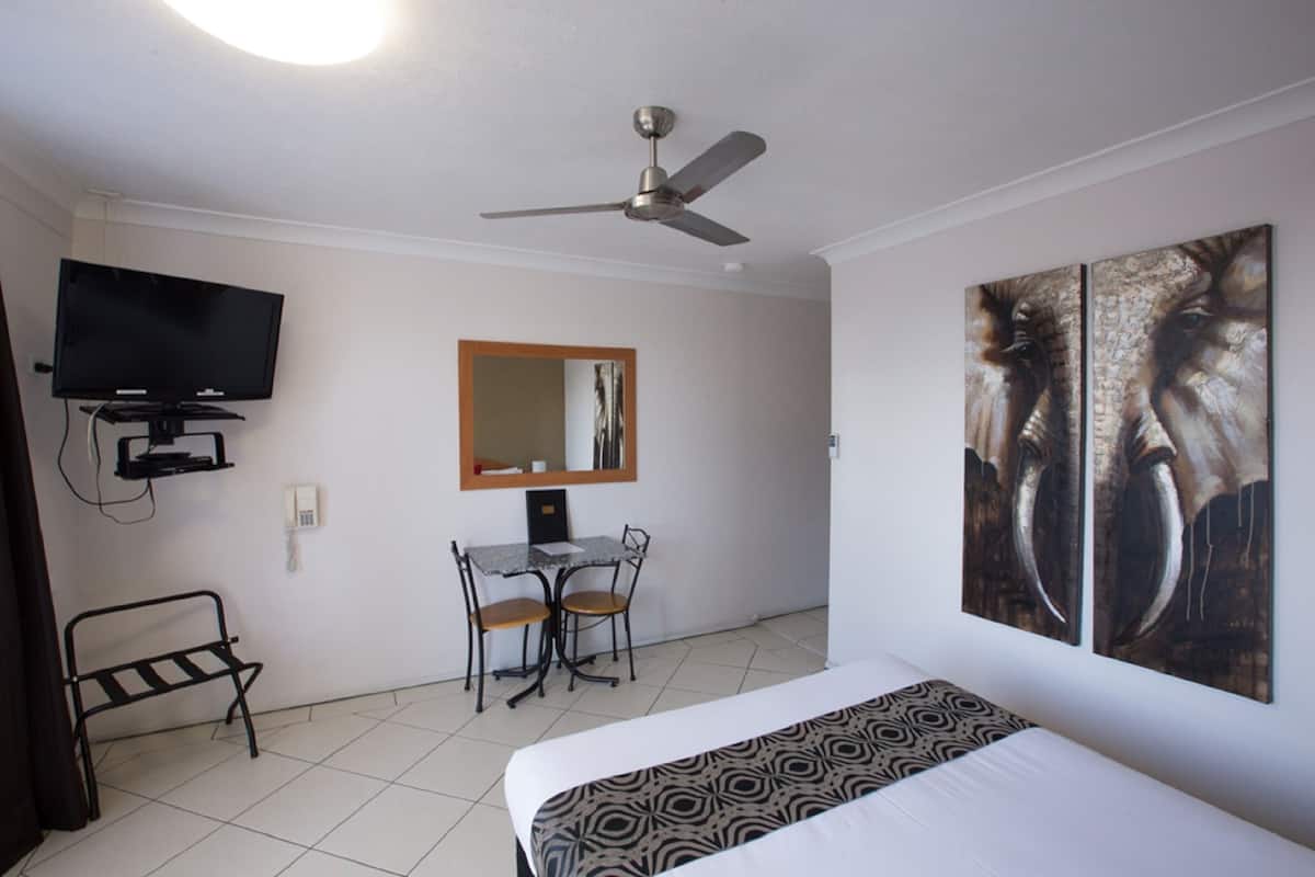 A hotel room with a bed, a desk with two chairs, a television, a ceiling fan, and artwork on the wall.