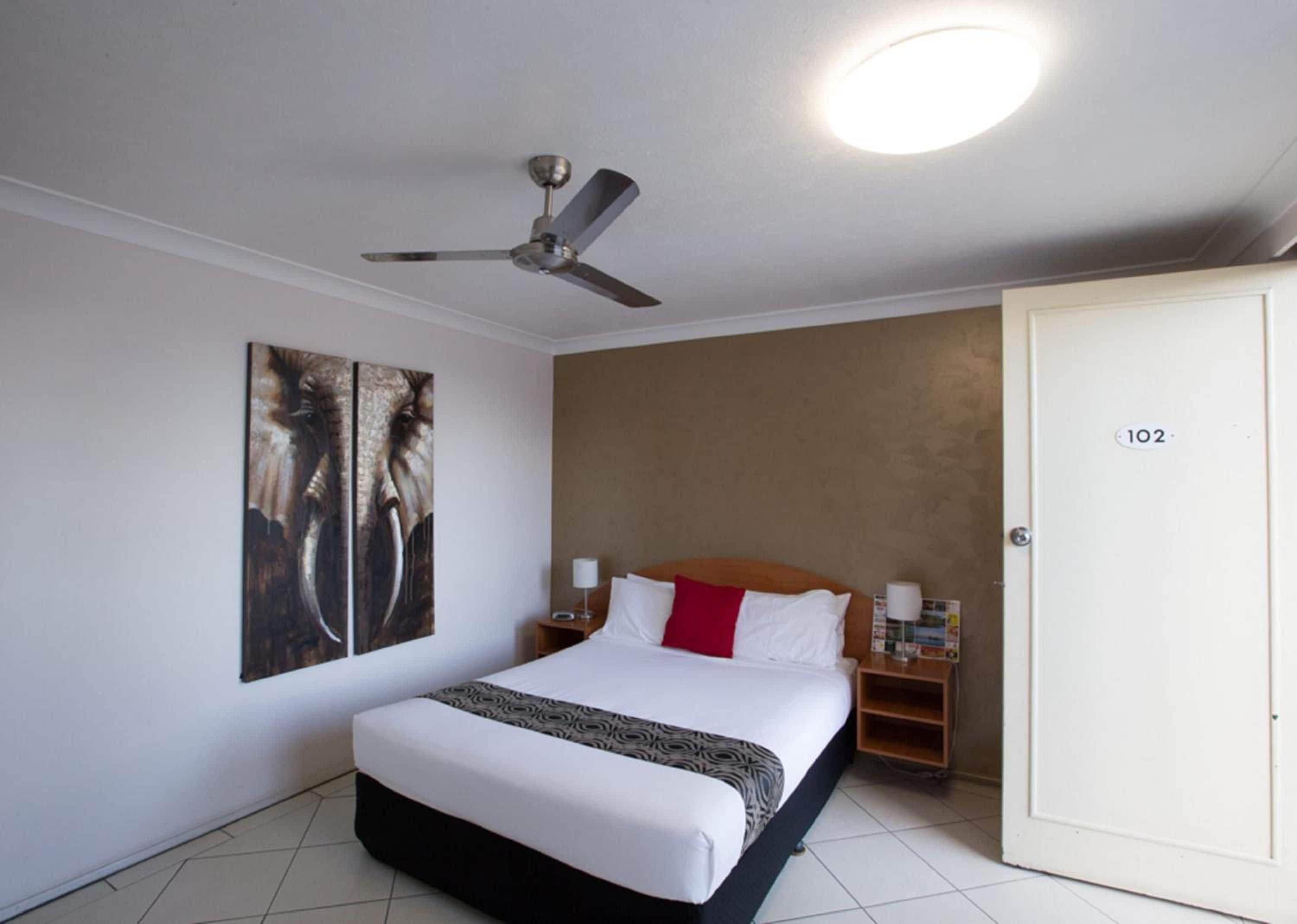 A hotel room with a bed, a ceiling fan, a wall-mounted artwork, a bedside table with a lamp, and a door with a number.