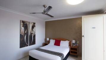 A hotel room with a bed, a ceiling fan, a wall-mounted artwork, a bedside table with a lamp, and a door with a number.
