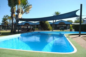 Ein Schwimmbecken mit einer umlaufenden Rasenfläche, Palmen und einem Gebäude mit blauem Dach im Hintergrund.