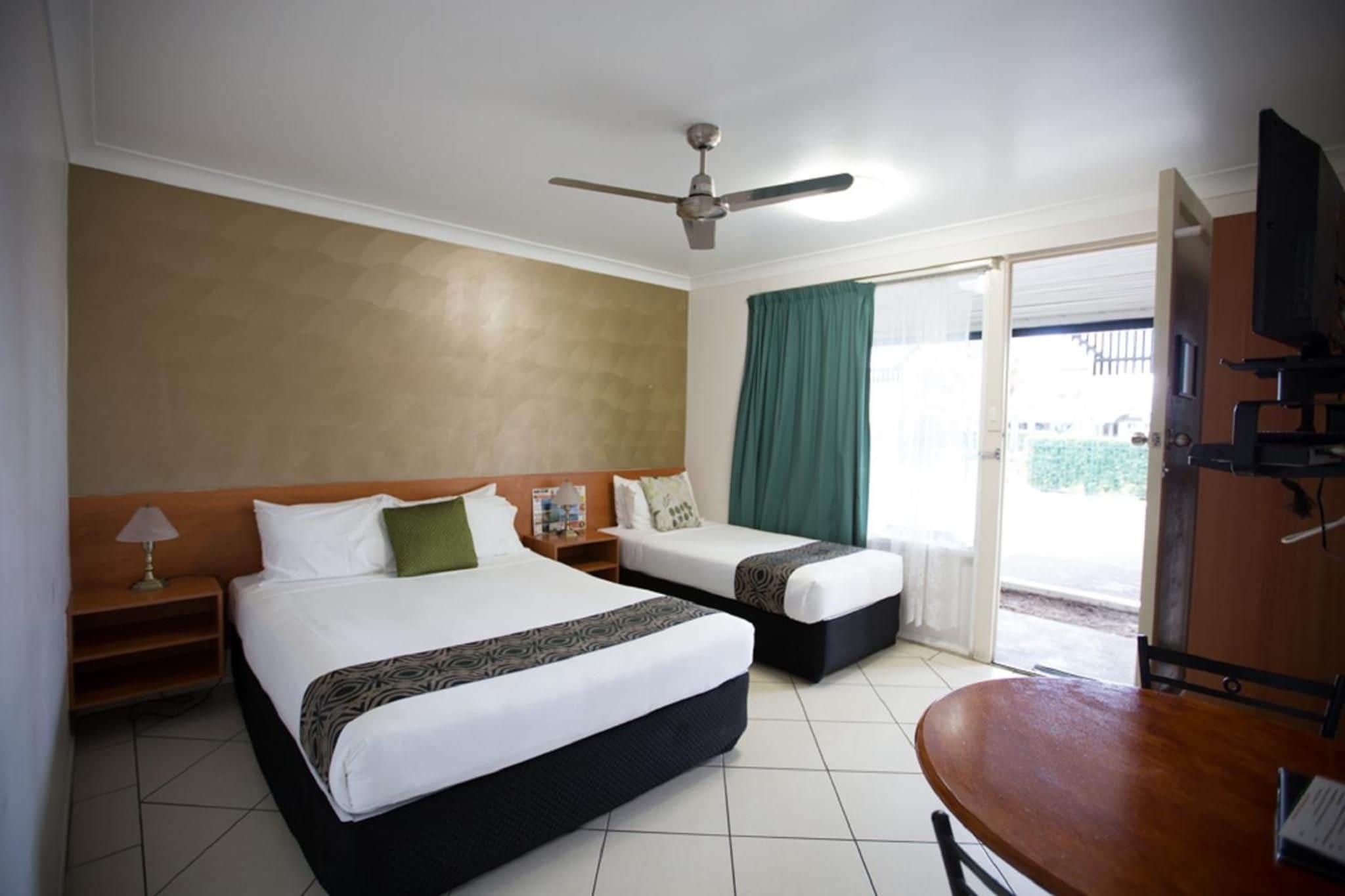Habitación de hotel con dos camas, ventilador de techo, televisor y puerta corrediza de vidrio al exterior.