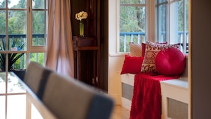 A room with a dining table, a bench with red cushions, and a window with a view of greenery. - Finlie (Hepburn)