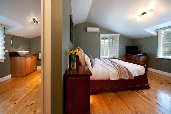 A bedroom with a wooden floor, a bed with a white comforter, a dresser, a vanity with a mirror, and a television on a stand.