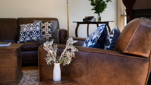 A brown leather sofa with patterned cushions, a white vase with dried flowers, and a small wooden table.