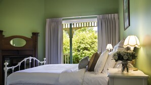 A bedroom with a large bed, a nightstand with lamps, a mirror, and a view of greenery outside.