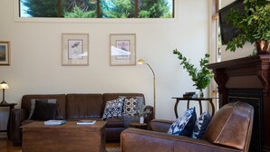 A living room with a brown leather sofa, a wooden coffee table, and a patterned rug. There are framed pictures on the wall, a floor lamp, and a potted plant.