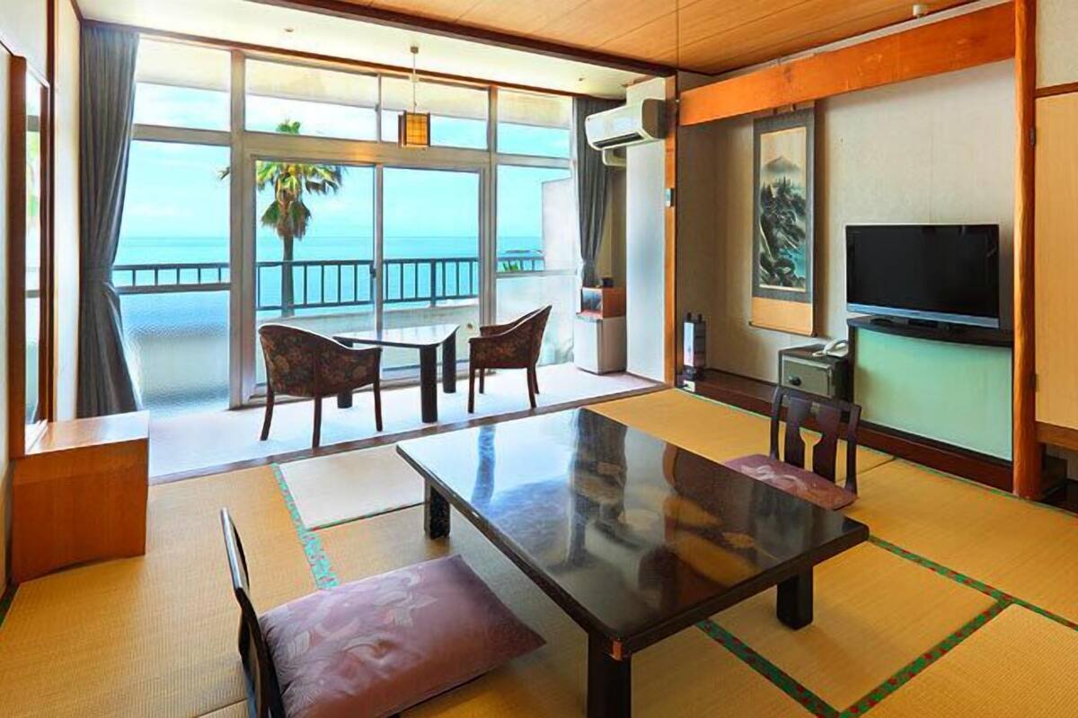 A room with a large table, chairs, a TV, and a view of the sea.