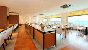 A modern hotel dining area with a long counter, white chairs, and large windows offering a view of the sea.