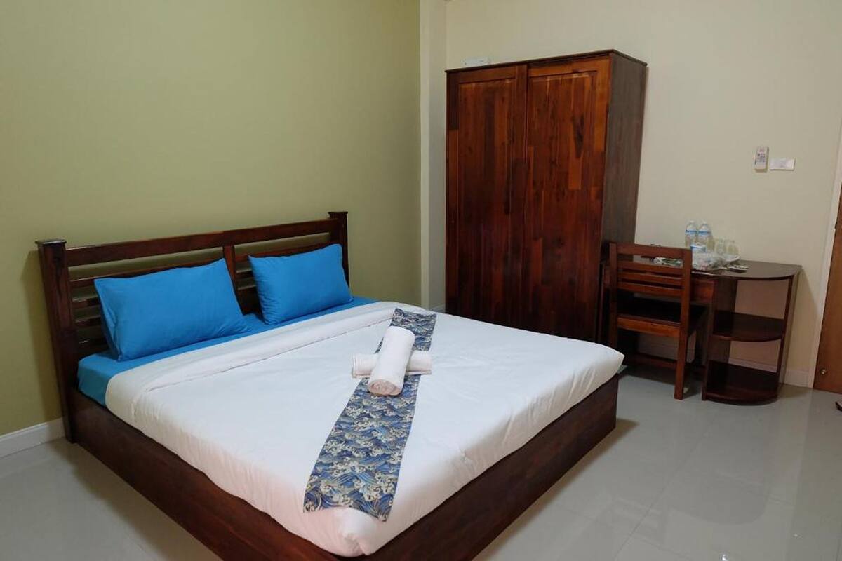 A neatly made bed with blue pillows, a wooden wardrobe, and a small desk with a chair.