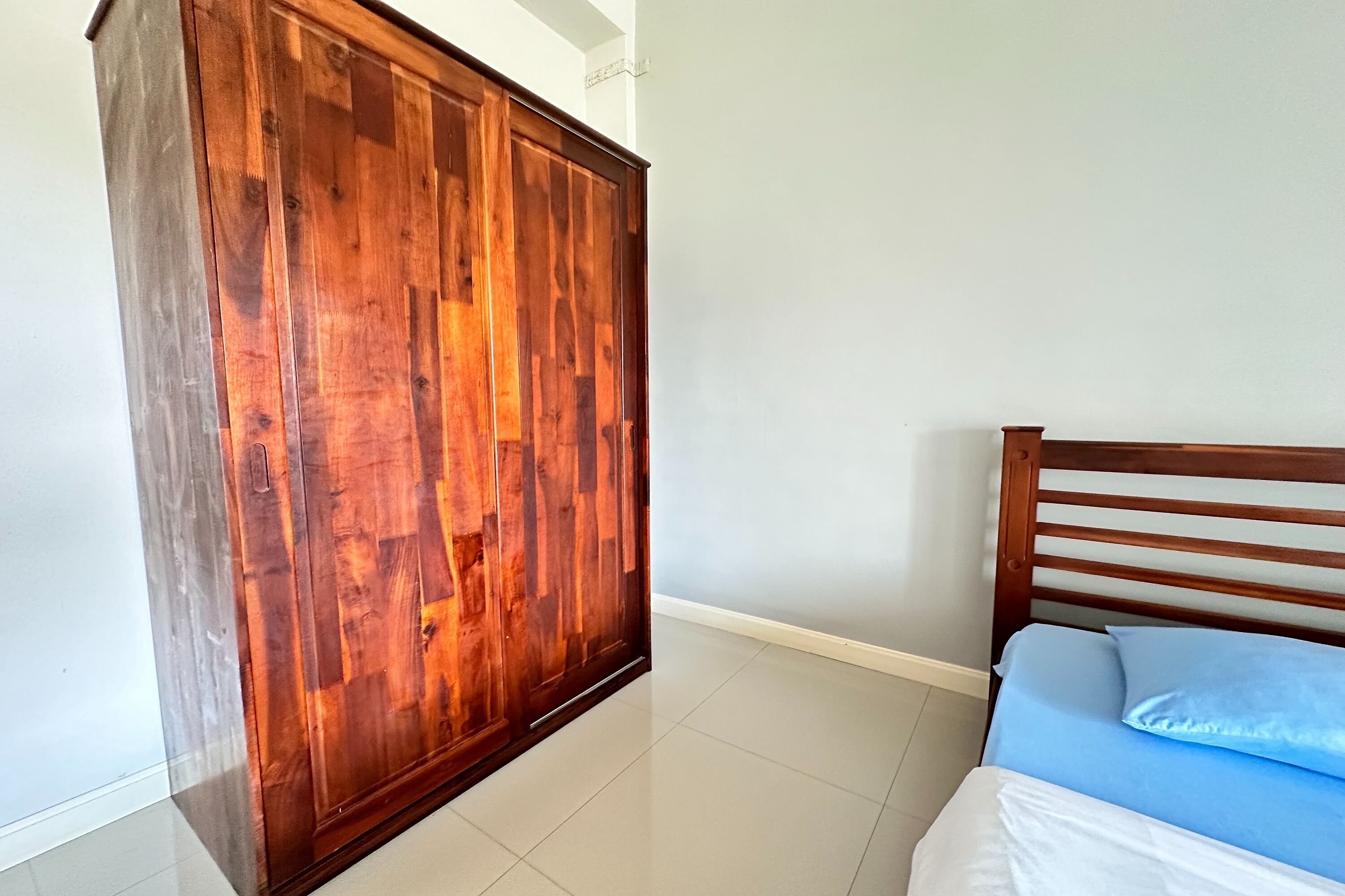 Un dormitorio con un armario de madera, una cama con ropa de cama azul y suelo de baldosas.